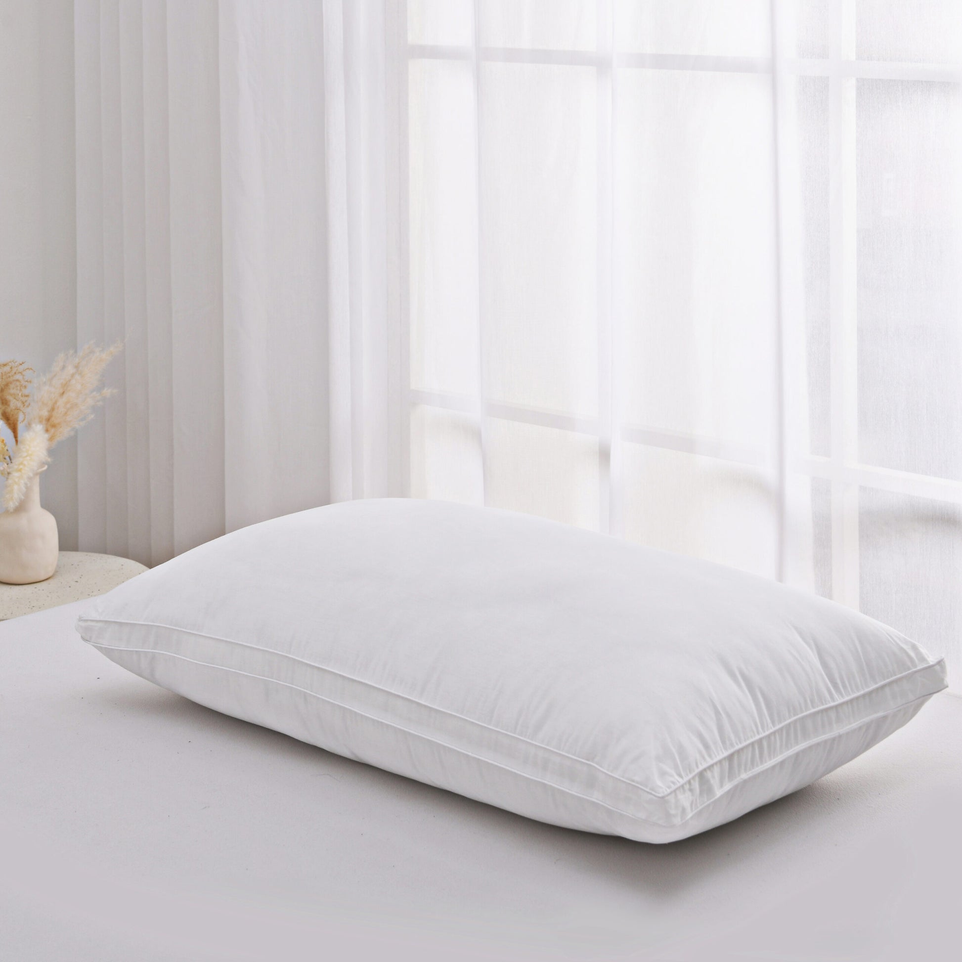 White microfibre pillow on a bed beside sheer curtains and dried pampas grass
